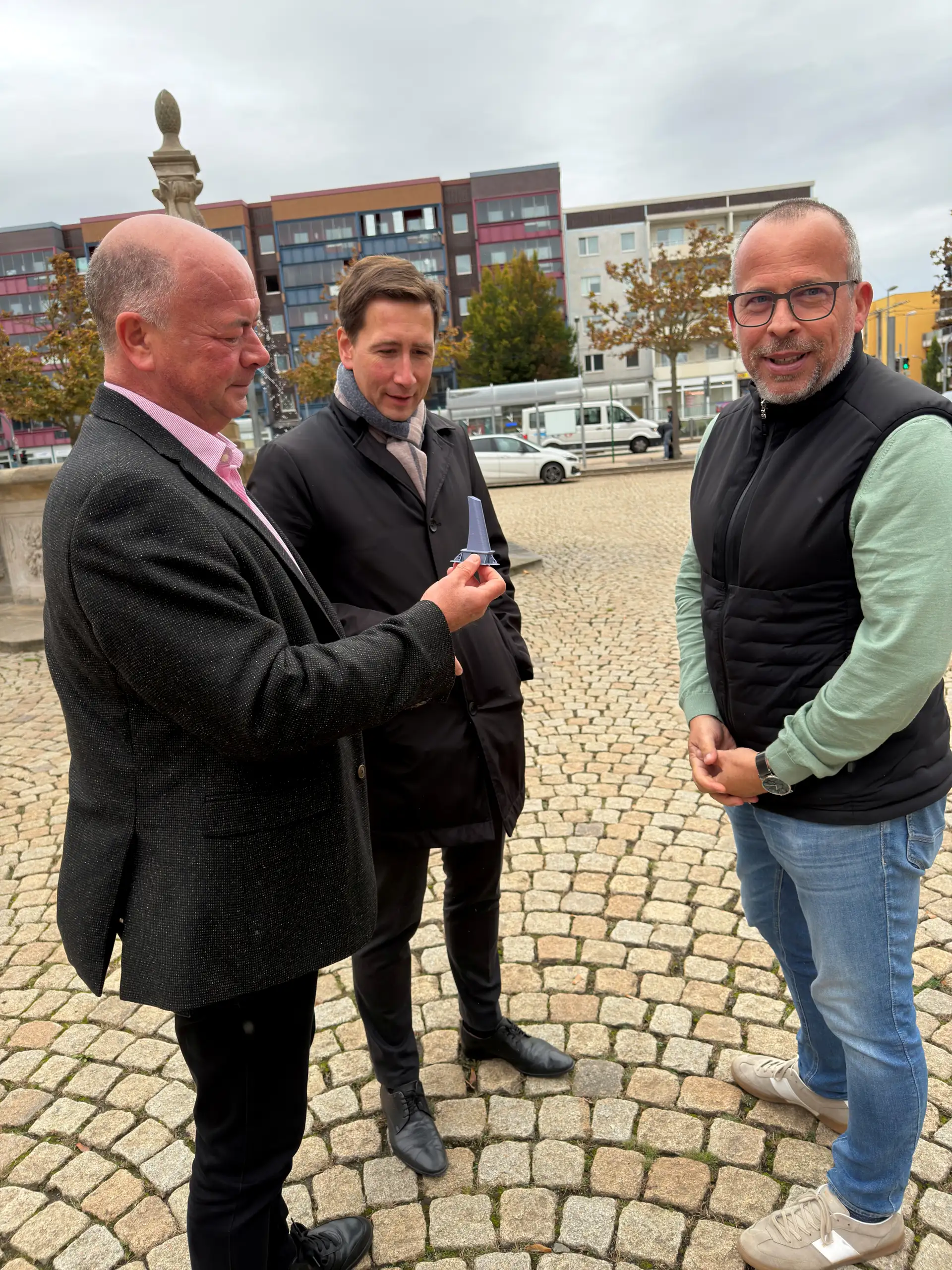 Drei Männer stehen auf dem Holzmarkt. Einer hält ein Modell eines Pollers, während die Gruppe die geplanten Sicherheitselemente bespricht.
